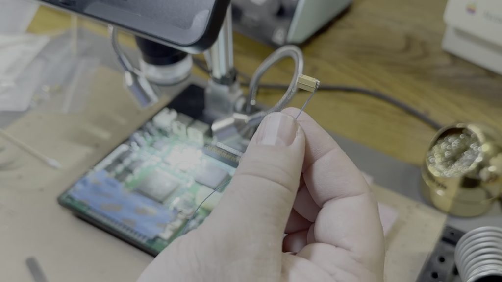 Capacitor and repair wire held above a Macintosh Classic II logic board in Michael Kamprath’s repair video